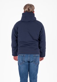 Giacca puffer blu navy con colletto alto e cappuccio, realizzata in materiale morbido, indossata sopra jeans in denim blu, con una vestibilità comoda.
