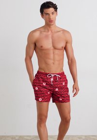 Rote Schwimmshorts mit einem wiederholten "HUGO"-Textmuster in Schwarz. Verfügt über einen elastischen Bund mit einer weißen Kordel und seitlichen Taschen.