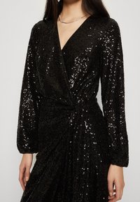 Svart paljettklänning med långa ärmar, v-ringning och knutdetalj i midjan. Tyget har en glänsande, strukturerad yta.