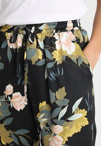 Pantalones negros con estampado floral que presentan una textura suave, una cintura elástica con cordón y vibrantes patrones de flores y hojas en rosa, amarillo y verde.