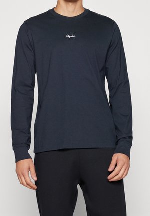 Mann trägt ein schlichtes dunkles Langarmshirt mit kleinem weißem "Rapha" Logo auf der Brust und dunkle Hose, steht vor einem schlichten Hintergrund.