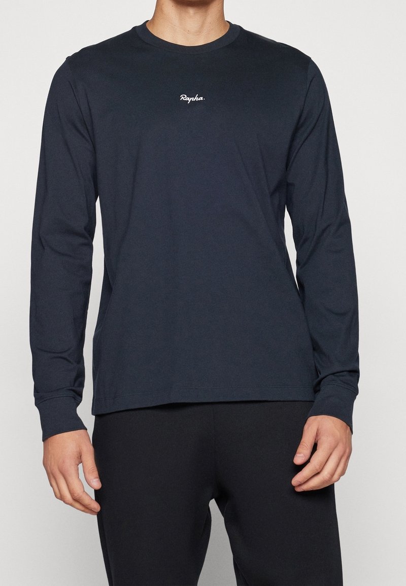 Homme portant une chemise à manches longues sombre unie avec un petit logo blanc "Rapha" sur la poitrine et un pantalon foncé, debout devant un fond uni.
