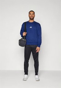 Φούτερ σε ναυτικό μπλε με λευκό λογότυπο, συνδυασμένο με μαύρο παντελόνι με λευκές ρίγες. Το μοντέλο κρατά μια μικρή μαύρη τσάντα και φορά λευκά αθλητικά παπούτσια.