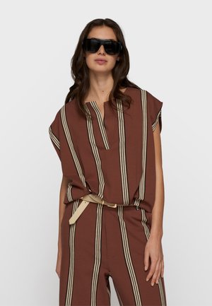 Kobieta w za dużych czarnych okularach przeciwsłonecznych i brązowym stroju z pionowymi beżowymi paskami oraz beżowym paskiem, stojąca na białym tle.