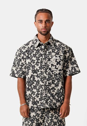 Hombre con camisa de manga corta y pantalones cortos a juego con patrones florales en blanco y negro, de pie contra un fondo blanco liso.