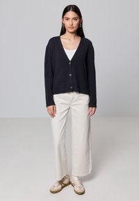 Granatowy sweter z dzianiny zapinany na pięć guzików, z dekoltem w serek, w zestawieniu z jasnobeżowymi spodniami typu wide-leg i kremowymi sneakersami. Gładka tekstura, swobodny krój.