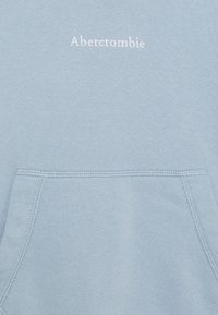 Světle modrá mikina s přední kapsou a bílým vyšitým logem "Abercrombie" poblíž výstřihu. Měkký bavlněný materiál.
