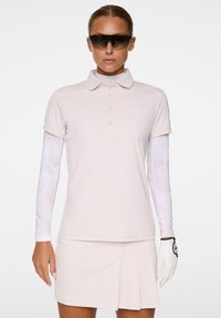 Jasnoróżowa koszulka polo z małym logo, krótkie rękawy, zestawiona z plisowaną spódnicą. Modelka ma na sobie okulary przeciwsłoneczne i białą rękawiczkę.