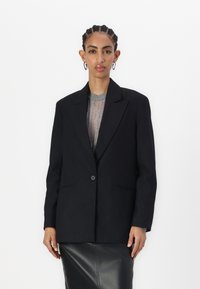 Blazer nero a righe con un singolo bottone, rever a punta e due tasche frontali. Indossato sopra un top grigio a trama e una gonna di pelle.