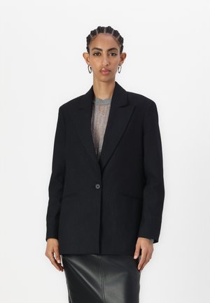Blazer nero a righe con un singolo bottone, rever a punta e due tasche frontali. Indossato sopra un top grigio a trama e una gonna di pelle.