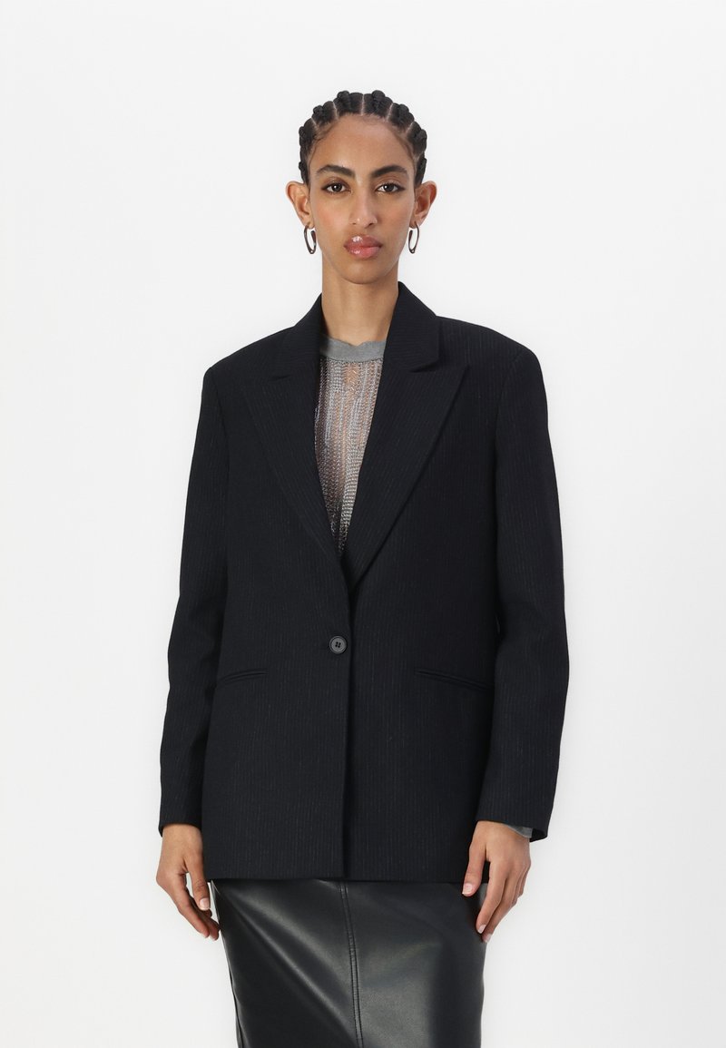 Blazer nero a righe con un singolo bottone, rever a punta e due tasche frontali. Indossato sopra un top grigio a trama e una gonna di pelle.