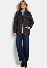 Vrouw in een zwarte gewatteerde jas, blauwe trui, donkerblauwe spijkerbroek en zwarte enkellaarsjes, staande tegen een witte achtergrond.