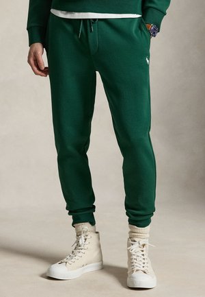 Άτομο με σκούρο πράσινο φόρμα παντελόνι με ρύθμιση στο τελείωμα, κρεμ ψηλά αθλητικά παπούτσια και μπεζ κάλτσες, στέκεται μπροστά σε απλό φόντο.