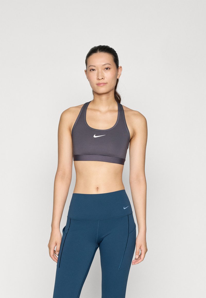 Nike sporta krūšturis tumši pelēkā krāsā ar noapaļotu kakla izgriezumu, sporta muguras dizainu un balto logo. Saskaņots ar augsto jostasvietu tirkīza krāsas leģingiem ar kabatām.