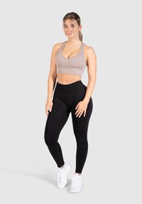 Sujeción deportiva en color taupe claro con textura acanalada, combinada con leggings negros de cintura alta, complementada con zapatillas blancas, sobre un fondo neutro.