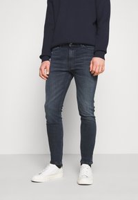 Mörkblå slim fit-jeans med en mjuk denimtextur, utrustade med framfickor och bältesöglor, ihop med vita sneakers och en marinblå sweatshirt.