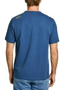 Mėlyna medvilninė marškinėliai trumpomis rankovėmis, apvaliu kaklu ir mažu auksiniu logotipu ant nugaros, derinami su džinsais. Minimalistinis dizainas.