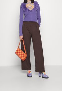 Femme portant un cardigan à col duveteux violet, un pantalon large marron, des chaussures compensées lavande, des chaussettes fleuries colorées, tenant un sac à main à carreaux orange.