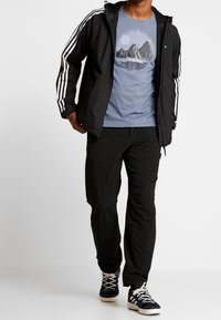 Svart lättviktsjacka med vita ränder, blå grafisk t-shirt med bergdesign, svarta byxor och blå skor med vita sulor.