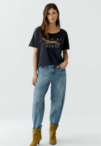 Short-sleeve, navy t-shirt with "WILD AT HEART" graphic in gold and white, paired with light blue, loose-fit jeans and tan ankle boots.