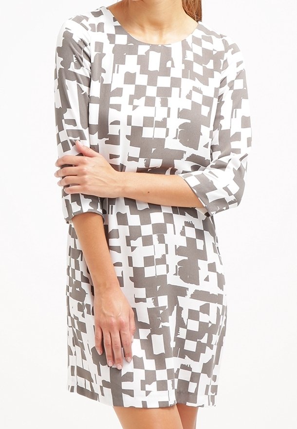 Femme portant une robe blanche avec des motifs carrés abstraits gris et des manches trois-quarts, debout les bras croisés devant.