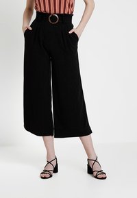 Black wide-leg trousers with a high waist, featuring a brown circular belt buckle, and two side pockets. Worn with black strappy heels.