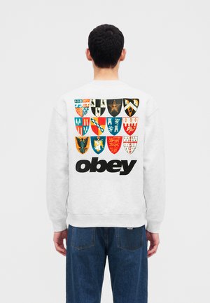 Uomo con felpa bianca con disegni colorati di scudi medievali e testo nero in grassetto "obey" sulla schiena, indossa jeans blu, di spalle.