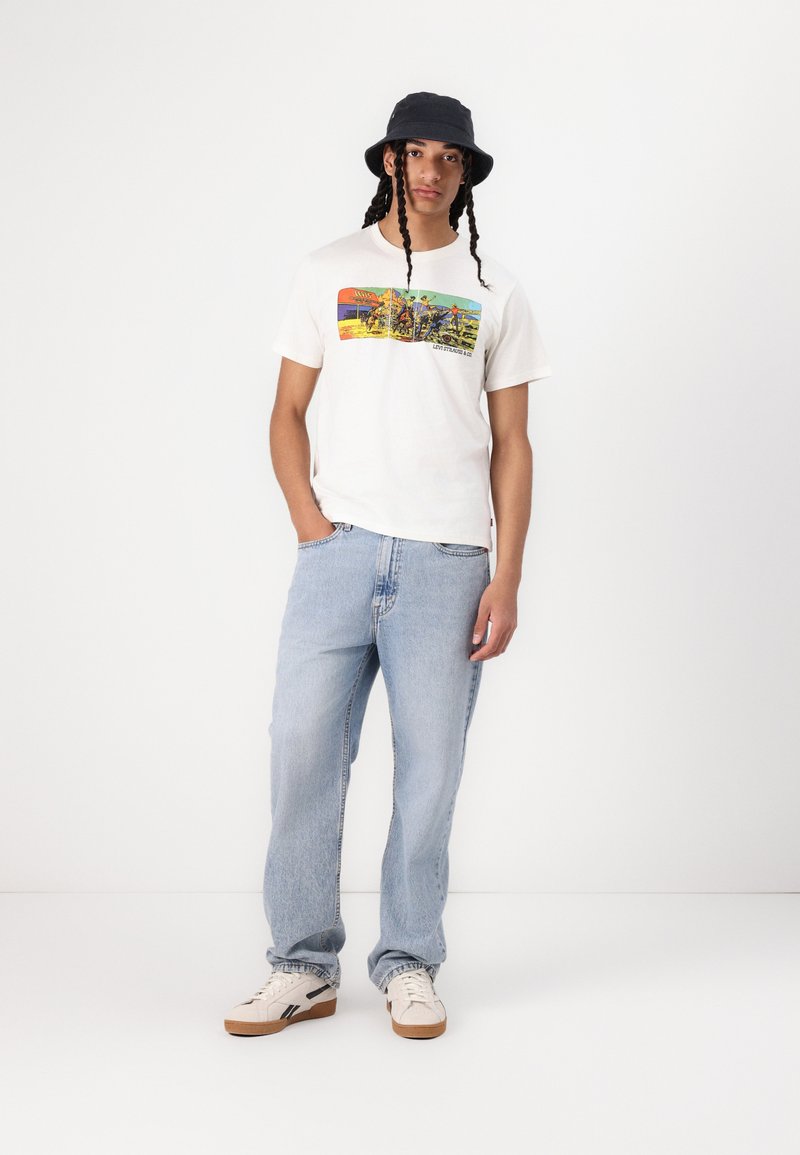 Weißes Baumwoll-T-Shirt mit einem bunten Grafikdruck auf der Vorderseite, kombiniert mit hellblauen Jeans und einem schwarzen Bucket Hat. Weiße Sneakers runden den Look ab.