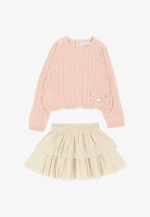 Maglione rosa a manica lunga lavorato a trecce con un piccolo fiocco e charm, abbinato a una gonna plissettata a strati color crema.