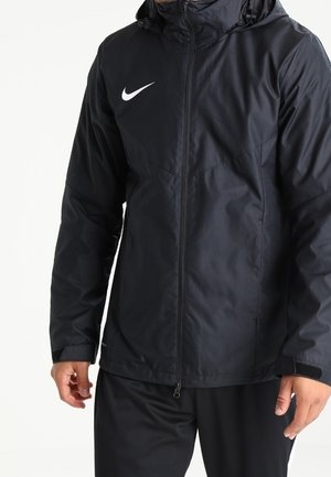 Homme portant une veste à capuche noire Nike avec une fermeture éclair à l'avant et un pantalon noir, debout devant un fond blanc uni.