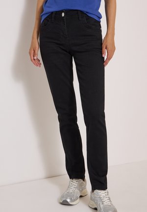 Persona che indossa pantaloni neri aderenti, una camicia blu e scarpe sportive argentate, in piedi su uno sfondo chiaro e uniforme.