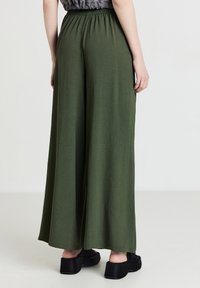 Person, der eine hoch taillierte, locker sitzende olivgrüne weit geschnittene Hose und schwarze Plateau-Schuhe trägt, steht vor einem einfarbigen hellen Hintergrund.