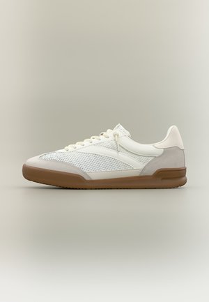 Zapatilla baja blanca y gris claro con paneles de malla y ante, cordones blancos y suela de goma marrón sobre un fondo neutro.