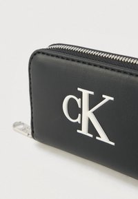 Μαύρο πορτοφόλι με φερμουάρ από υφή τεχνητής δερματίνης, με εμφανές λευκό λογότυπο "CK" και ασημένια εξαρτήματα φερμουάρ.