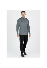 Maglia atletica grigia a maniche lunghe con mezza zip e dettagli neri, abbinata a pantaloni neri aderenti con dettagli a bottoni lungo il lato.