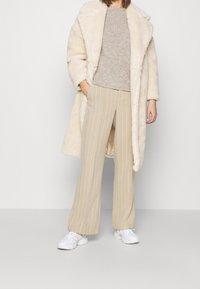 Femme portant un pantalon beige à rayures larges, un pull en tricot gris, un manteau en fausse fourrure couleur crème, et des baskets blanches, debout avec une main dans la poche.
