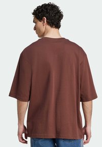 Barna pamut, oversized póló rövid ujjakkal, kerek nyakkal és sima textúrával, a ruházat hátulnézeti kilátását mutatja.