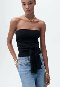 Top nero a coste senza spalline con lati arricciati, dettaglio con cintura annodata, abbinato a jeans in denim blu chiaro. Accenti di bracciale e collana dorati.