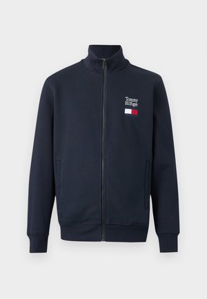 Tumši zila zip-up jaka ar stāvošu apkakli, sānu kabatām un nelielu Tommy Hilfiger logotipu un karogu kreisajā krūtīs.