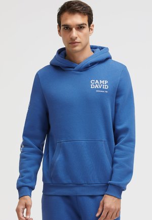Junger Mann trägt einen blauen Camp David Hoodie mit Fronttasche und Schriftzug auf der Brust, steht vor einem einfarbigen hellen Hintergrund.