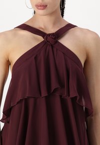 Burgundska halter top haljina s volanima, s čvorom na izrezu. Izrađena od laganog, lepršavog materijala.