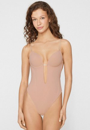 Vrouw met krullend haar, gekleed in een naakte kleurige bodysuit met diepe V-hals en dunne bandjes, staand tegen een effen achtergrond.