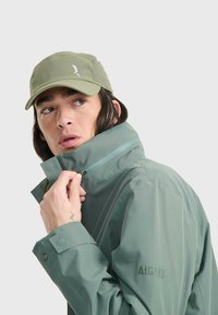 Veste imperméable verte avec col montant et fermeture éclair, arborant le logo AIGLE, associée à une casquette vert clair avec des détails en mesh.