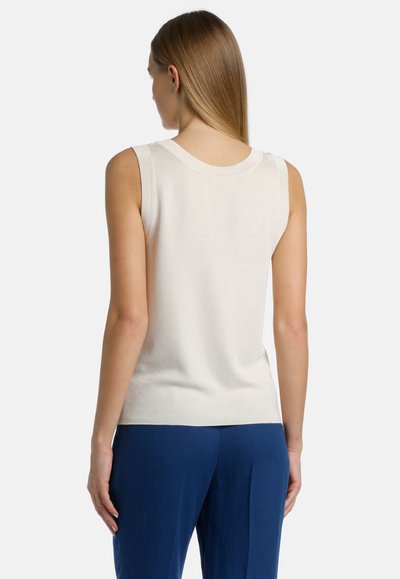 Top de punto sin mangas en color crema con escote redondo; textura suave y diseño ajustado, combinado con pantalones azul oscuro de corte ajustado visibles en las caderas.