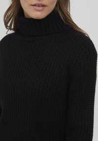 Pull à col roulé en tricot noir avec une texture côtelée. Doté d'un col haut et d'une coupe décontractée, mettant en avant un matériau cosy et épais.