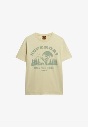 Camiseta beige de manga corta con gráfico verde de montañas, árboles, un pájaro y texto "SUPERDRY Pikes Peak Council Dist." en el frente.