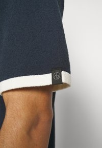 Gros plan d'une manche courte bleu foncé avec bordure blanche et une petite étiquette noire avec un logo blanc circulaire sur le bras d'une personne.