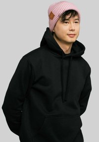 Czarna bluza z kapturem i kieszenią kangurką; jasnoróżowa czapka z prążkowanej dzianiny z brązowym skórzanym akcentem z boku; neutralne tło.