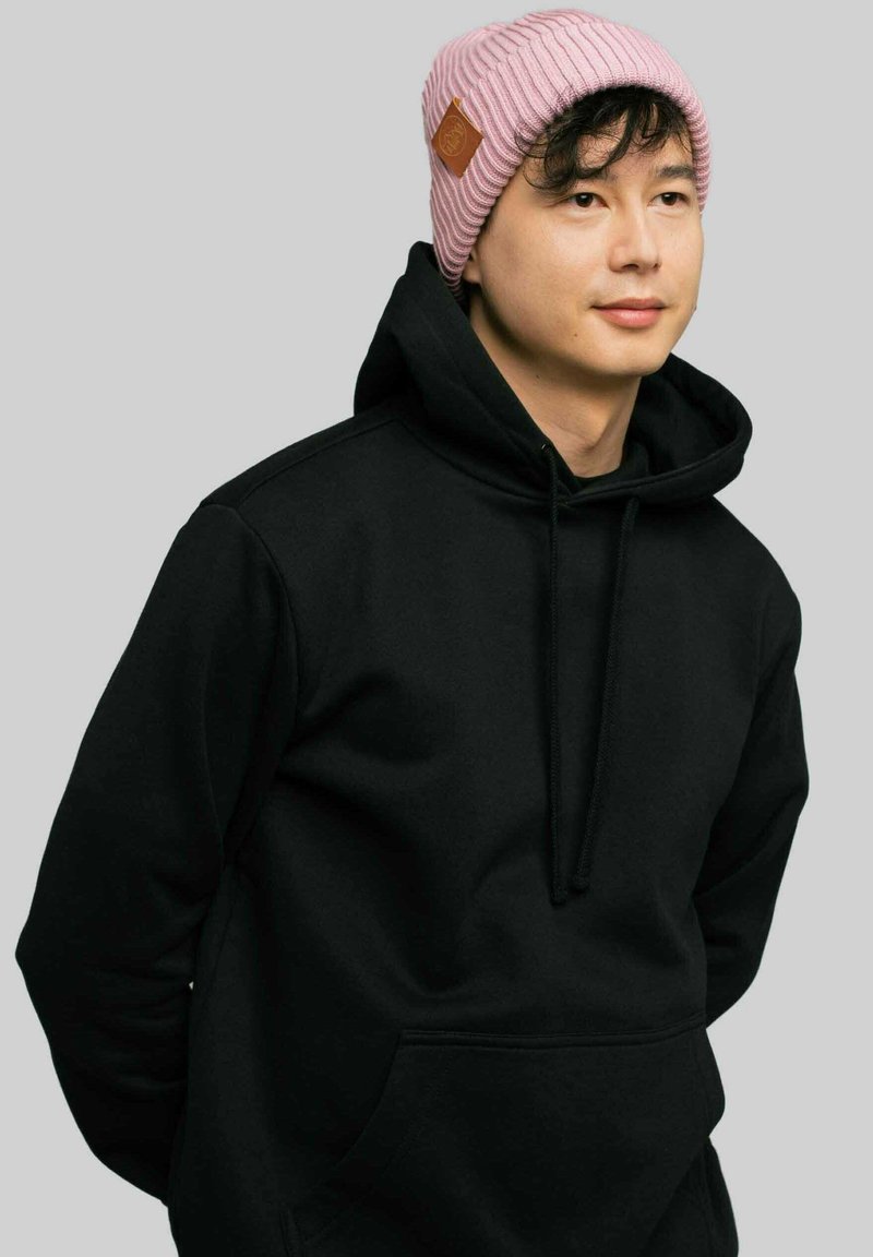 Czarna bluza z kapturem i kieszenią kangurką; jasnoróżowa czapka z prążkowanej dzianiny z brązowym skórzanym akcentem z boku; neutralne tło.