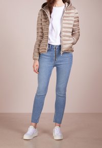 Helle beige Pufferjacke mit Reißverschluss und Kapuze, kombiniert mit hellblauen, enganliegenden Jeans und weißen Turnschuhen. Glatte Textur und gestepptes Design.
